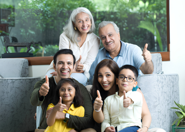 A three-generation Indian family sitting together in a living room and giving a thumbs-up gesture while smiling at the camera.